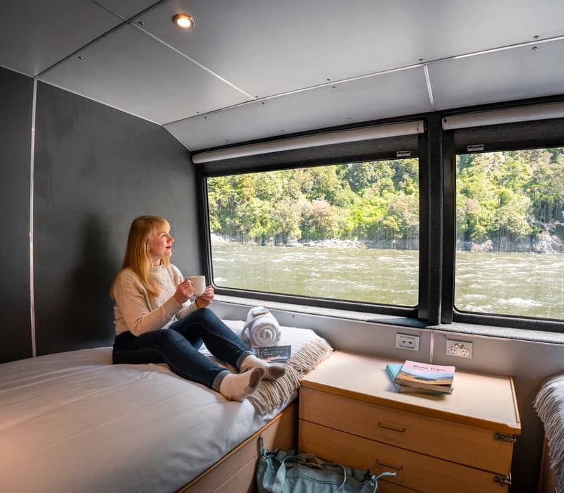 Woman sitting on bed with cup looking out window at river and forest in a cabin.