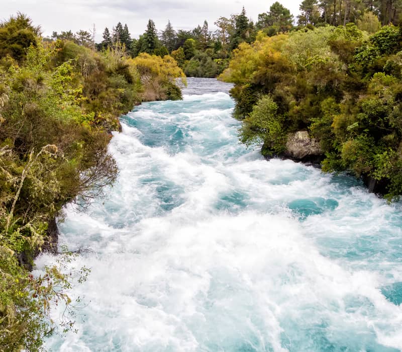 White and blue rapids of Huka Falls river surrounded by dense green forest under cloudy sky.