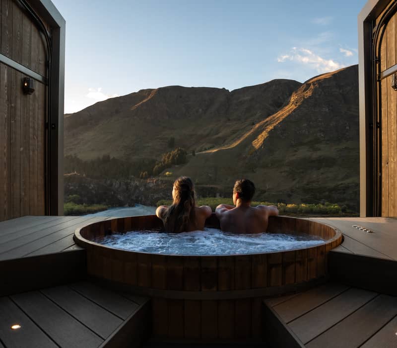Couple in wooden hot tub facing mountains and river at sunset