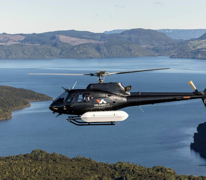 Black Volcanic Air helicopter flying over a blue lake with forested hills and mountains.