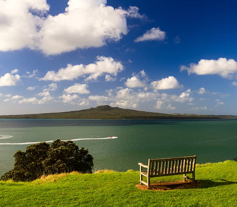 Bench on grassy hill overlooking water with island, boats, and cloudy blue sky.