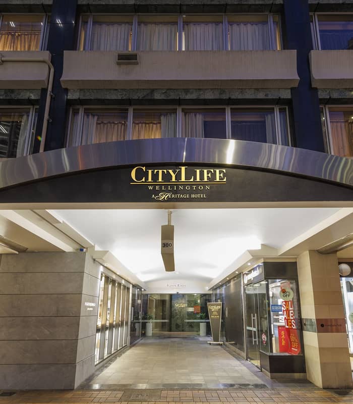 Illuminated hotel entrance to CityLife Wellington with shops and sale signs on either side at night.