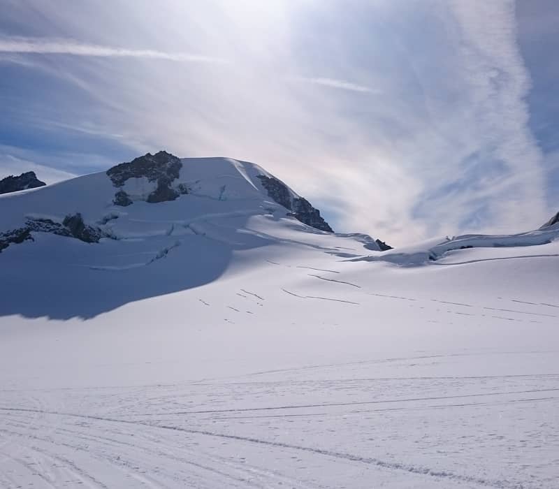 A vast, pristine snowfield on the upper reaches of Franz Josef Glacier, with deep crevasses visible on the mountain slopes under a bright sun and wispy clouds.