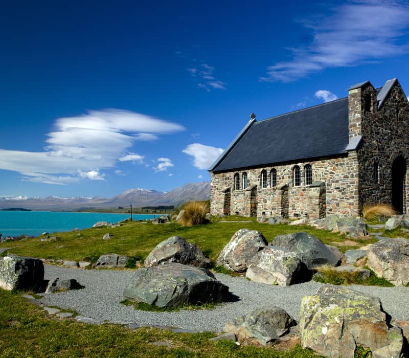 Stone church near turquoise lake with mountains and blue sky in the background.