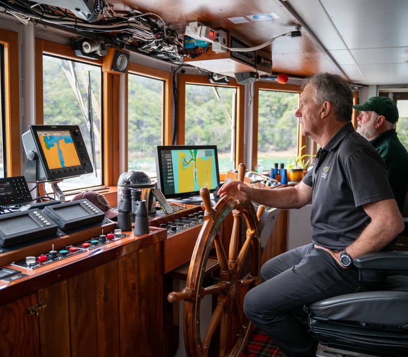 Skipper steering a cruise boat with passengers inside the cabin and navigation screens visible.