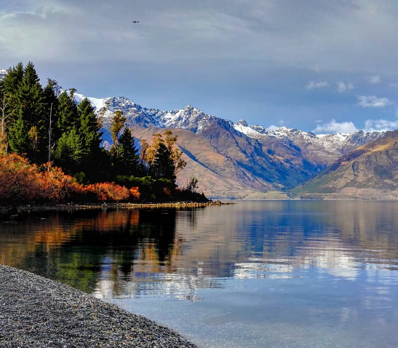 Lake with clear water reflecting snow-capped mountains and trees on a partly cloudy day.