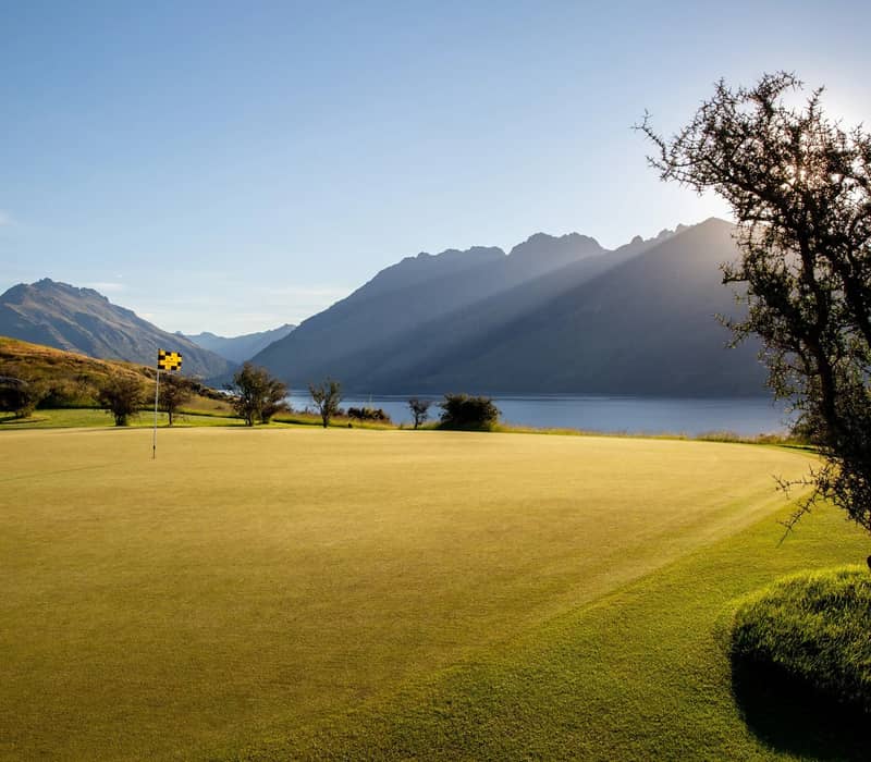 Golf green with flagstick near a lake, mountains in the background, and sunlight streaming through trees.