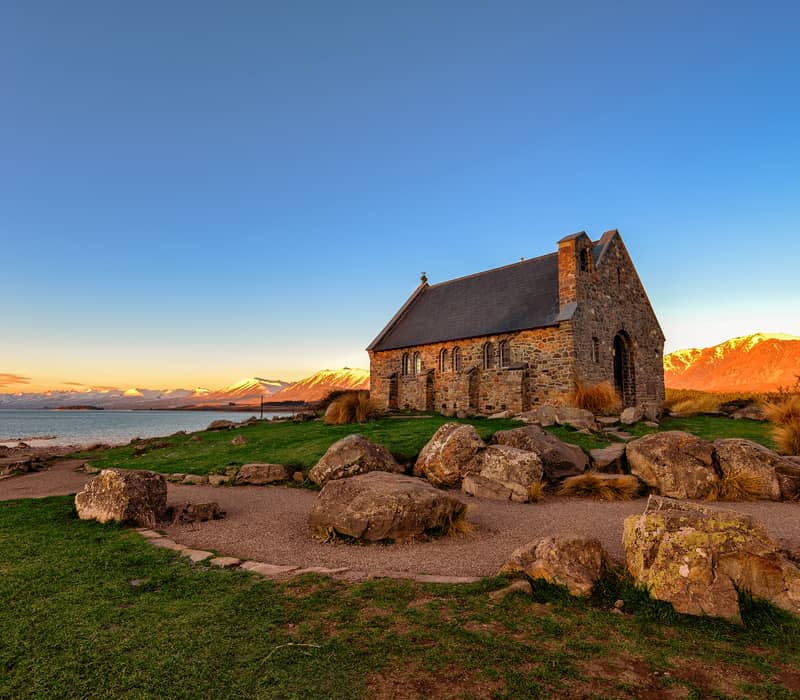 Stone church at sunset near a lake with mountains in the background.