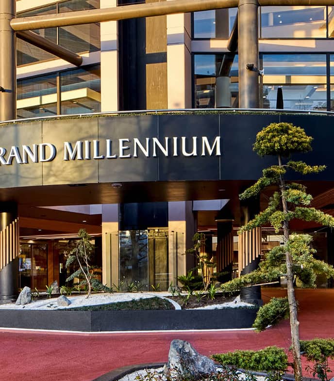 Modern entrance of Grand Millennium hotel with glass walls, columns, plants, and red carpet driveway.