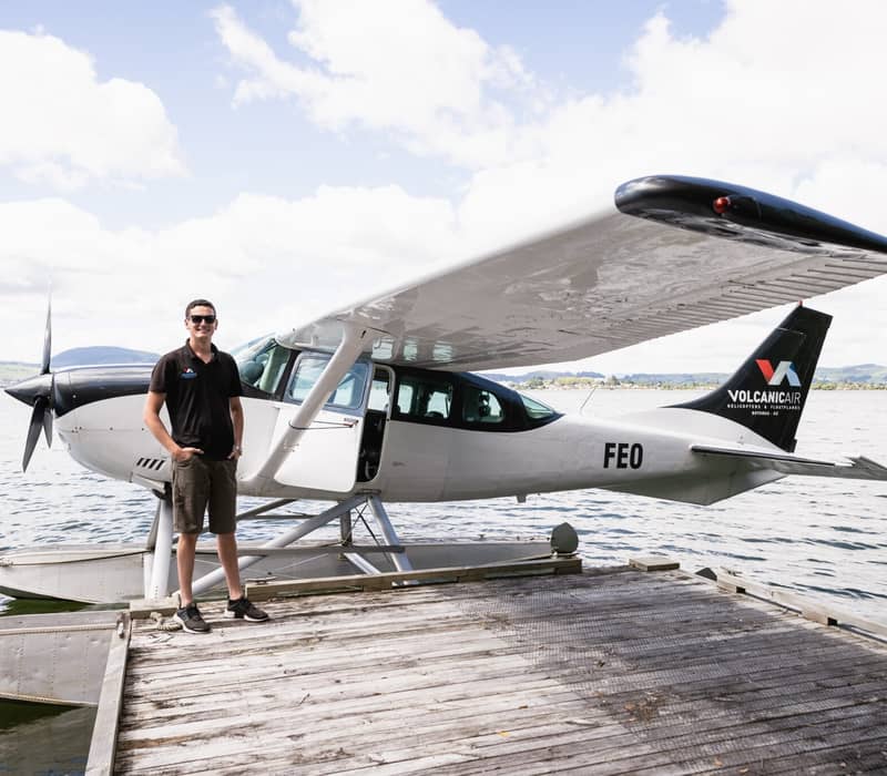 Pilot standing on dock beside a floatplane on a lake with mountains and clouds.