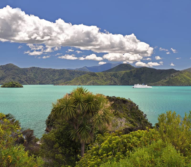 White ferry on turquoise bay water with green islands and hills, under blue sky with clouds.