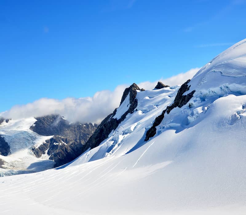 A bright, sunlit view of a steep, snow-covered mountain ridge with blue ice crevasses, set against a vibrant blue sky in the Southern Alps.