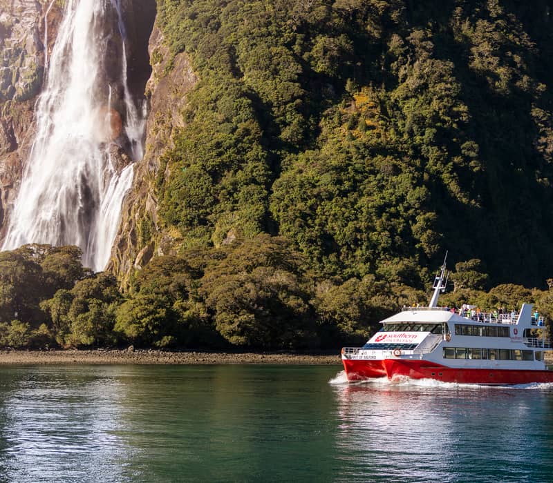 Red and white cruise boat near a waterfall and forested mountain in Milford Sound Credit: Southern Discoveries