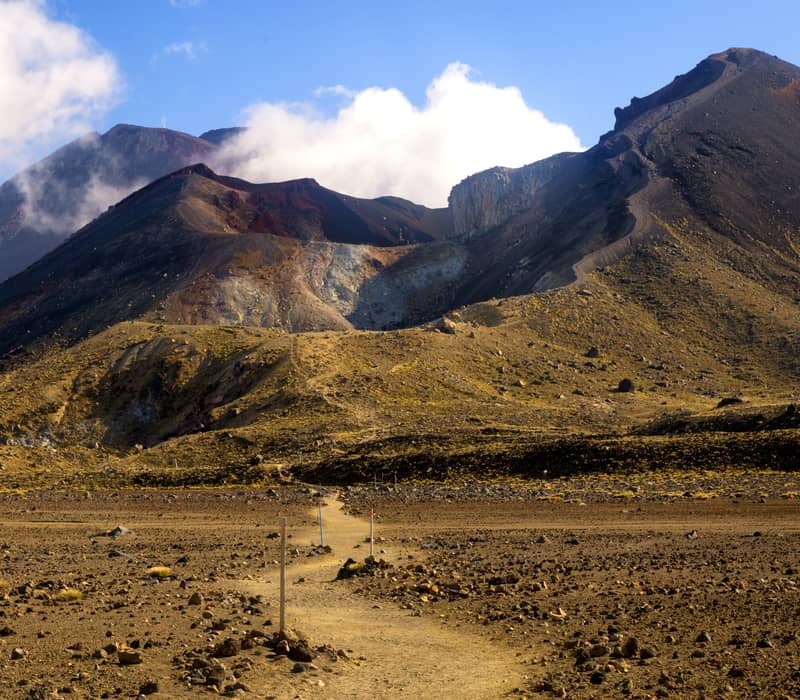 Volcanic mountain Mt Ngauruhoe with hiking trail in foreground under blue sky with clouds.