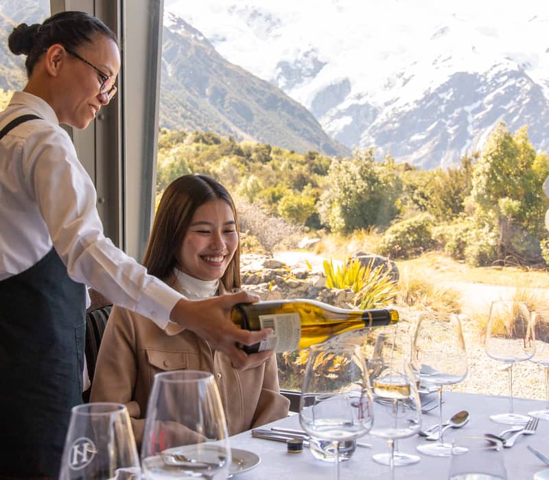 Waiter pouring white wine to a woman at a restaurant table with mountains in the background
