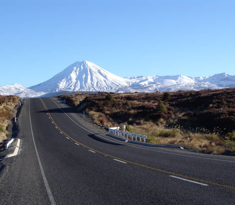 Paved road heading towards snow-covered Mount Ngauruhoe volcano under a clear blue sky.