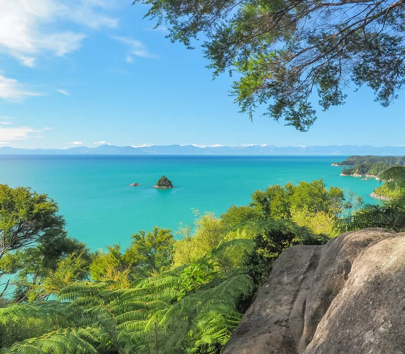 Looking through green trees at a small rocky island in a bright blue sea.
