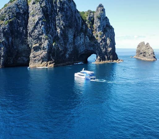 Boat sailing near rocky island with natural arch in blue ocean under clear sky.