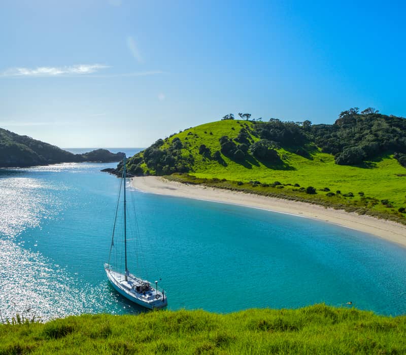 Bright blue sky with green verges, a curved sandy beach and the blue sea with a small boat in the centre 