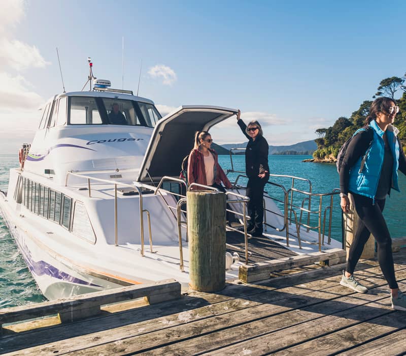 People getting off a white ferry boat onto a wooden dock with green trees and blue water in background.