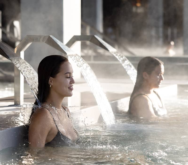 Women relaxing under hydrotherapy water jets in a spa pool with steam around.