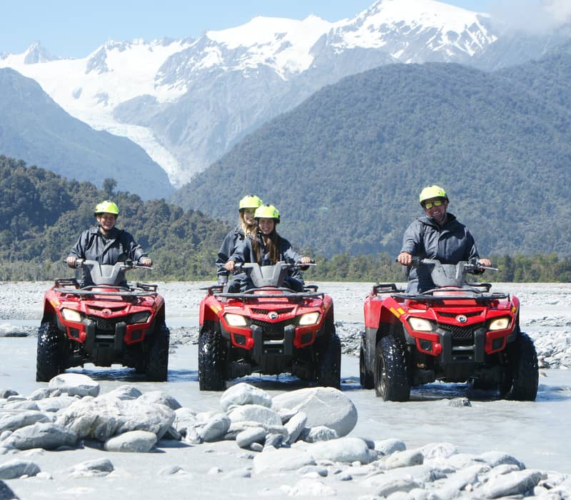 Four people riding red ATVs across a rocky riverbed with snowy mountains and glacier behind.