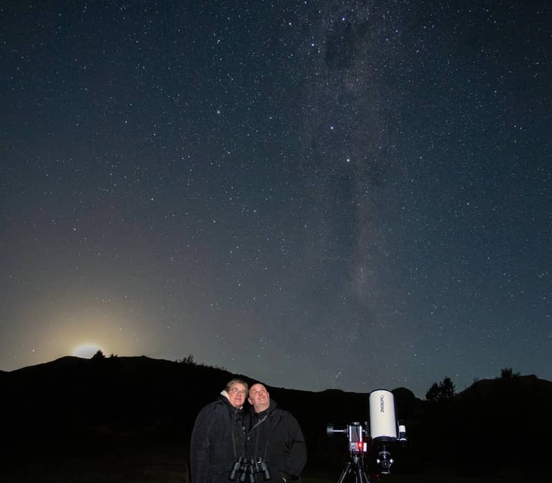 Couple stargazing with telescope under starry night sky and Milky Way over mountains.