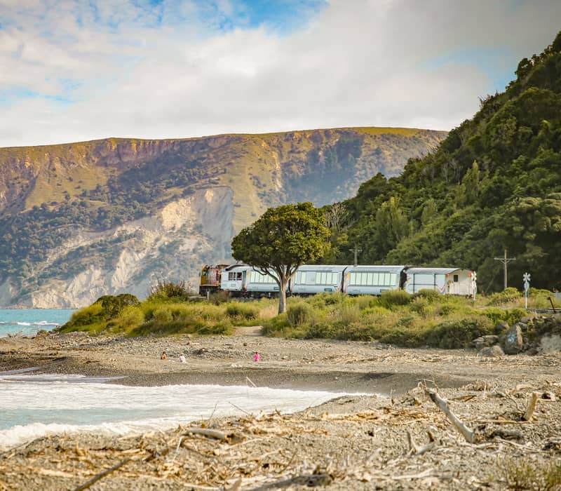 Train moving along beach coastline with mountains and ocean in the background.