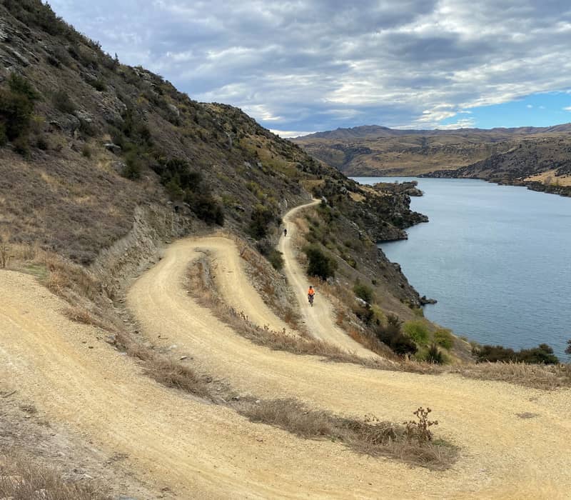 Cyclists ride along a winding dirt road on a hillside next to a lake under a cloudy sky.