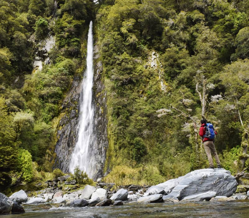 Hiker with backpack standing on rock looking at tall waterfall surrounded by dense forest.