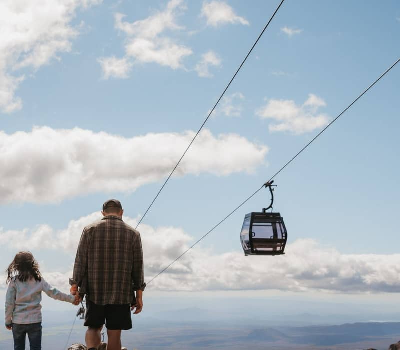 Adult and child holding hands hiking on rocky mountain trail near cable car under blue sky with clouds.