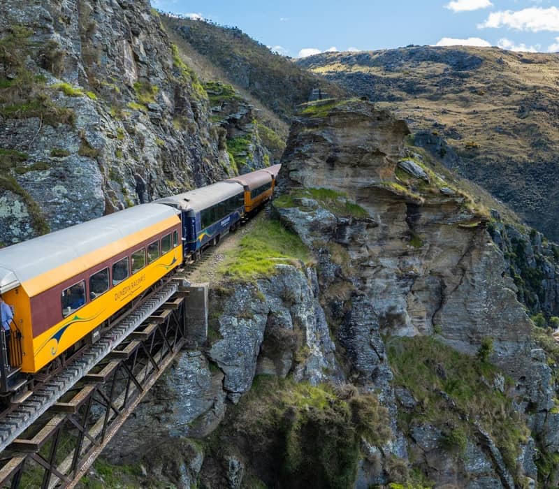 Passenger train on railway bridge crossing rocky gorge with river below and mountainous landscape.