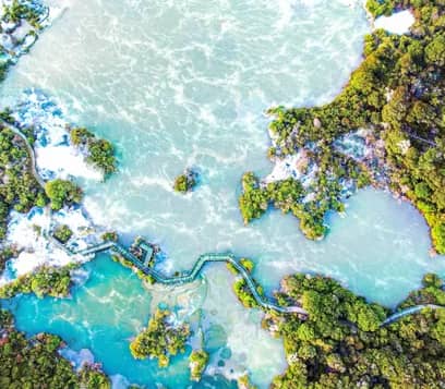 An aerial bird's-eye view of a winding wooden boardwalk crossing through vibrant turquoise and pale green geothermal pools surrounded by dense green native bush.