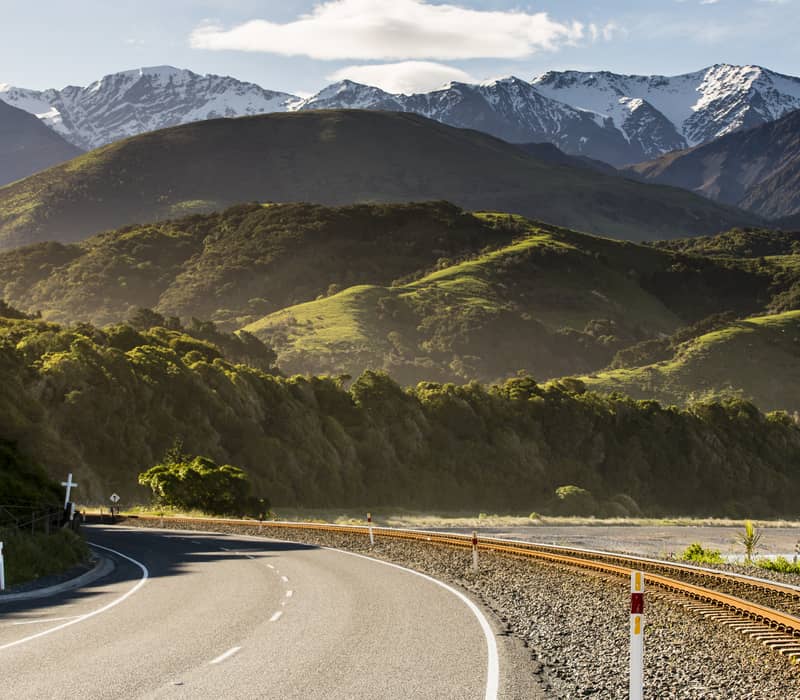 Road and railway track next to green hills with snowy mountains in the background.