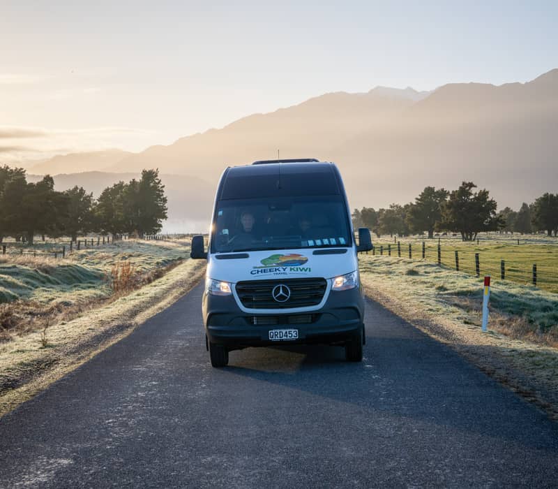 A travel van drives down a country road with mountains and trees in the background at sunrise.