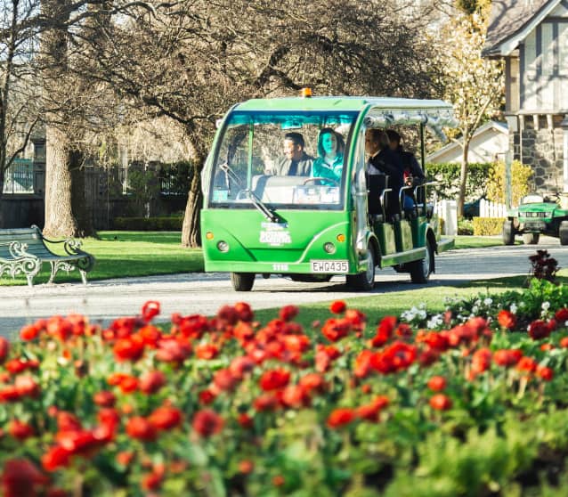 Green garden tour vehicle driving past red flowers with passengers inside on a sunny day.