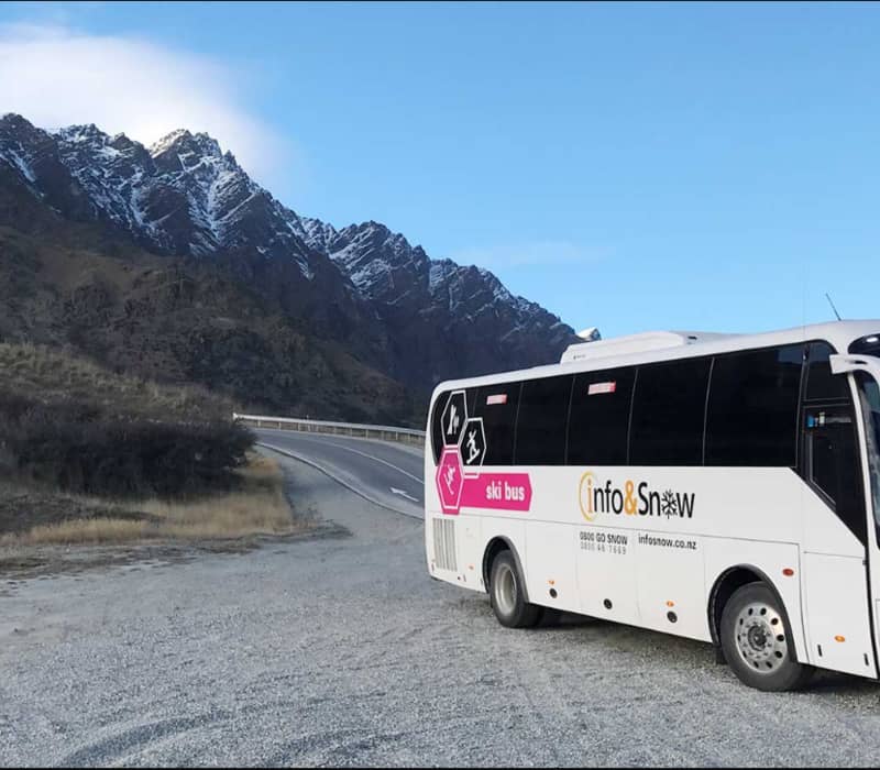 White ski bus parked on a gravel road with snow-covered mountains behind under a clear blue sky.