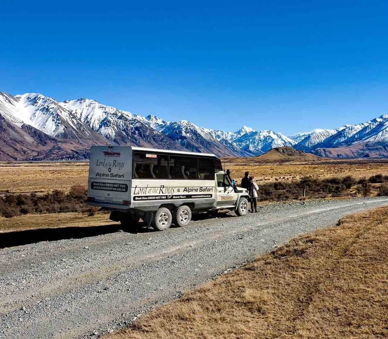 4x4 tour bus parked on a dirt road with people beside it and snow-covered mountains in the background under a clear sky.