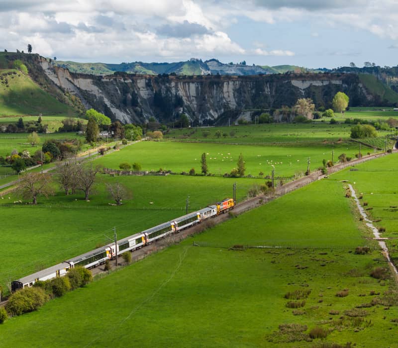 Northern Explorer train traveling on railway track through green farmland near Rangitikei Viaduct with hills behind.