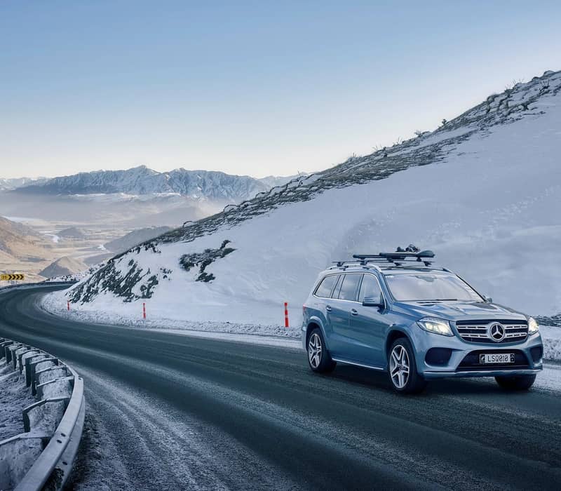 Blue SUV driving on snowy mountain road with snow-covered peaks in the distance under clear sky.