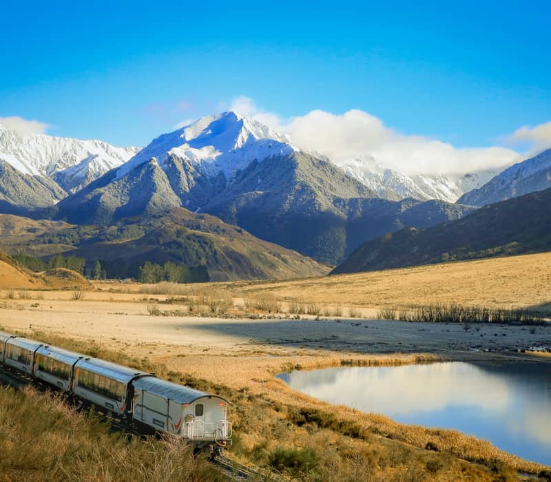 Train passing through valley with snow-covered mountains and lake reflecting sky and clouds
