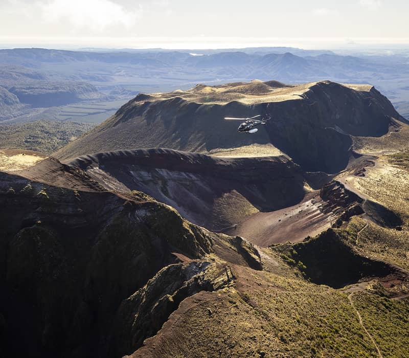 Helicopter flying over the crater of Mount Tarawera volcano with rugged terrain and distant mountains Credit: PB & DV Holdings Ltd T/A Volcanic Air