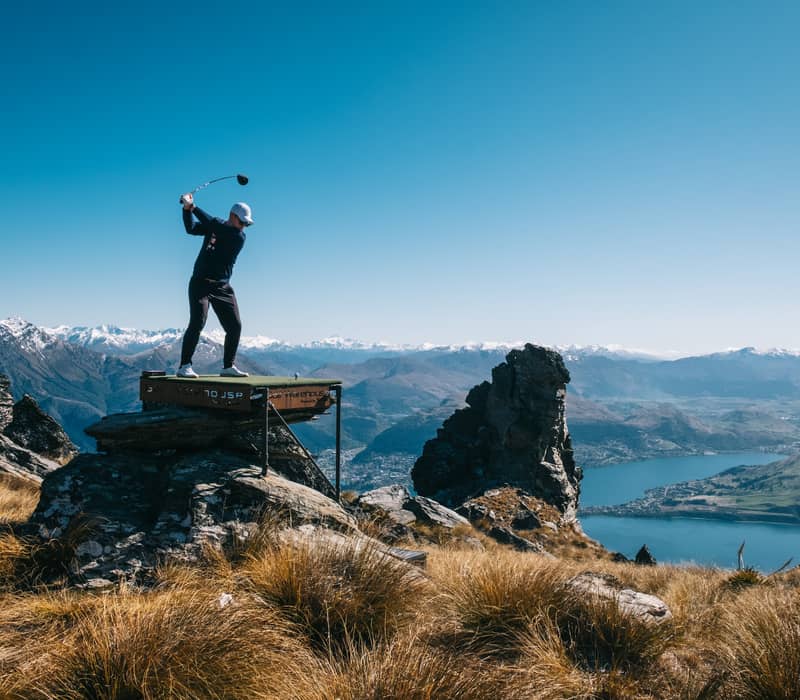 Golfer swinging club on elevated platform with mountains and lake in the background.