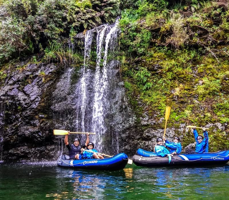 People kayaking in blue rafts near a waterfall with green forest and rocks.