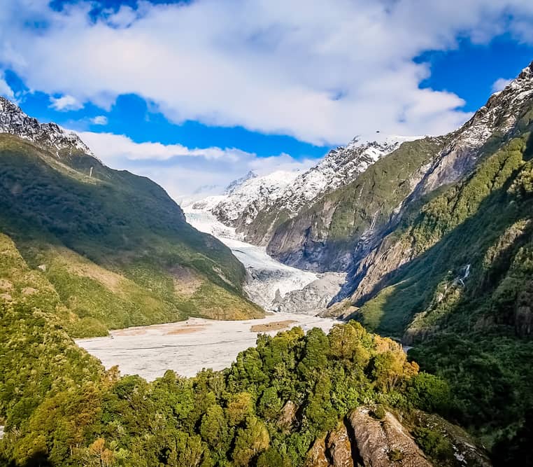 A wide valley view showing the white ice of Franz Josef Glacier descending between steep, green forest-covered mountains under a blue sky with white clouds.