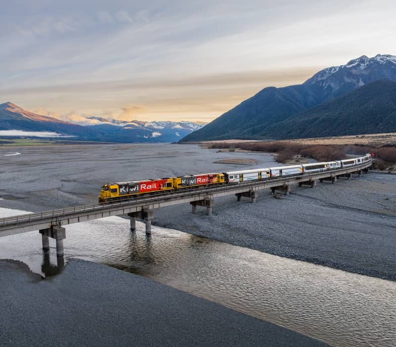 Aerial view of a KiwiRail train on a bridge over river with mountains and clouds at sunset.