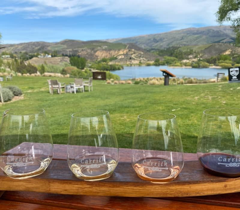 Four wine glasses with a wine flight in front of a lake, hills, and grassy field.