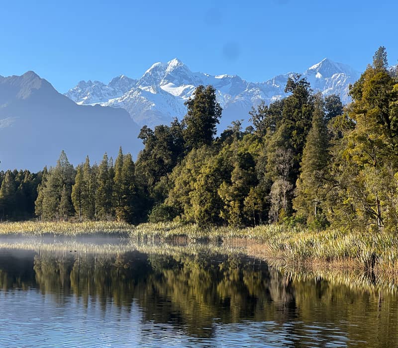 Calm lake with forest trees and snow-covered mountains reflected on water under clear sky.