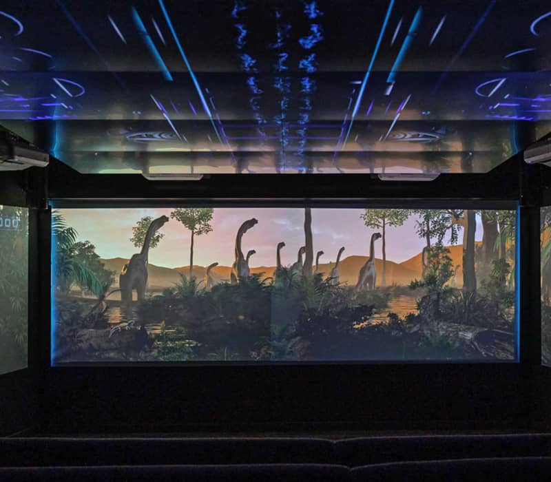 Dark projection room with screens showing animated dinosaurs and prehistoric landscape.