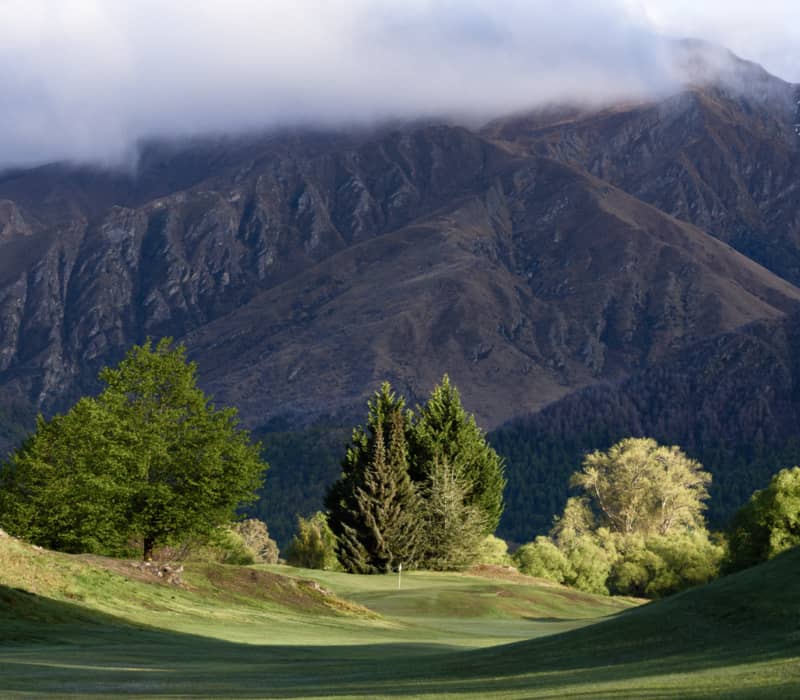 Green golf course with trees and mountains partially covered by clouds in the background.